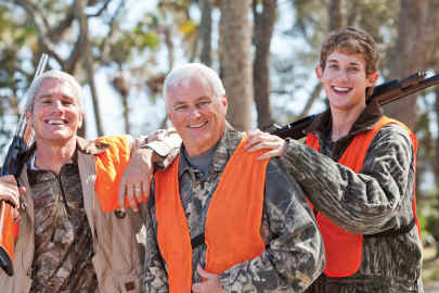Three cross-generational men smile in the woods in their hunting equipment