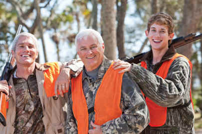 Three cross-generational men smile in the woods in their hunting equipment