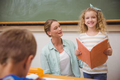 A school aged girl smiles after reading in front of her class next to her teacher, who looks proud of the girl's progress